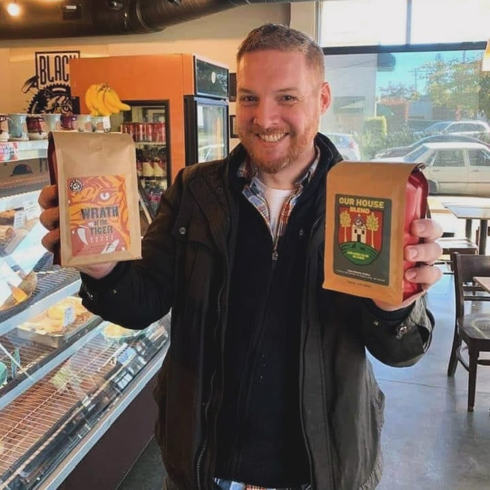 A smiling man holds up two bags of Jim & Patty's coffee in a cafe.