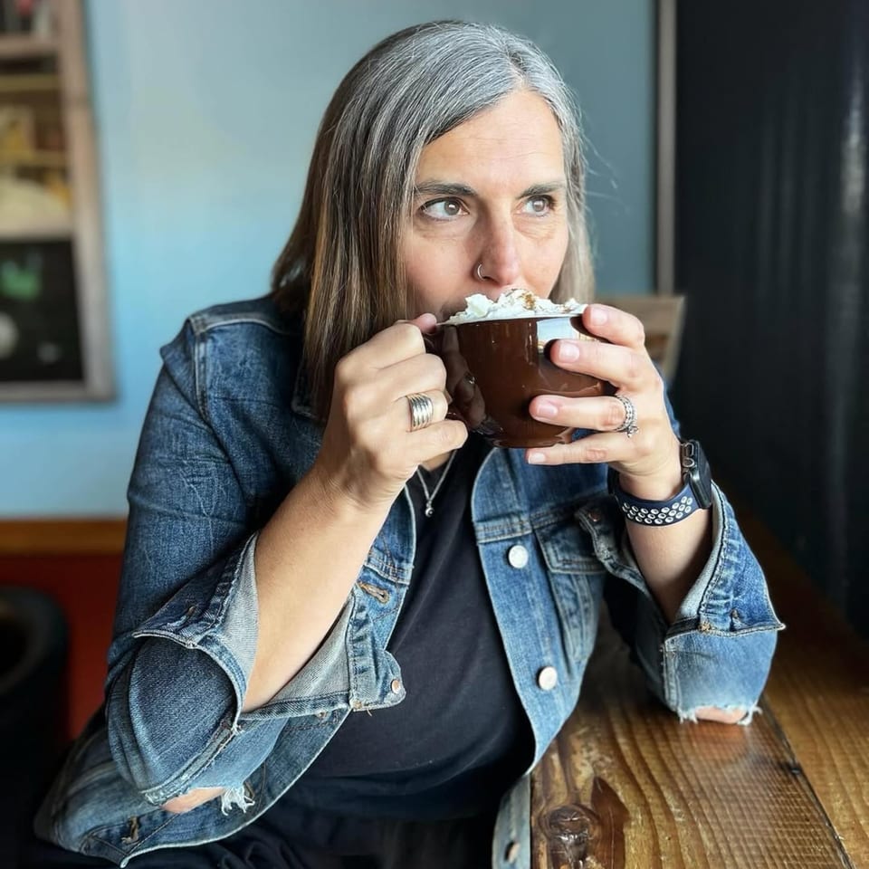 A woman with gray hair sips a coffee drink topped with whipped cream from a brown mug.