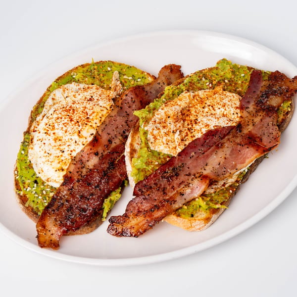 Two avocado toasts topped with fried eggs, paprika, and crispy bacon strips on a white plate.