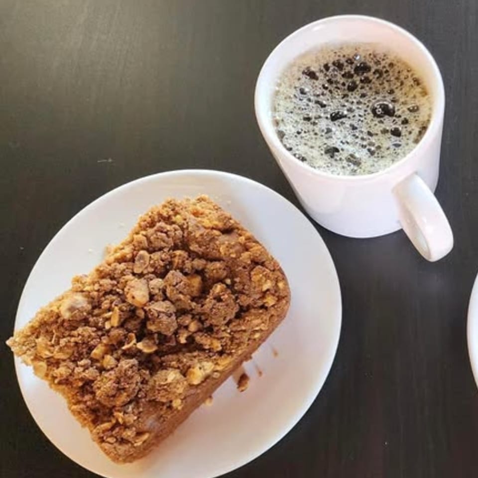 A coffee cake with crumble topping on a white plate next to a white mug of coffee.