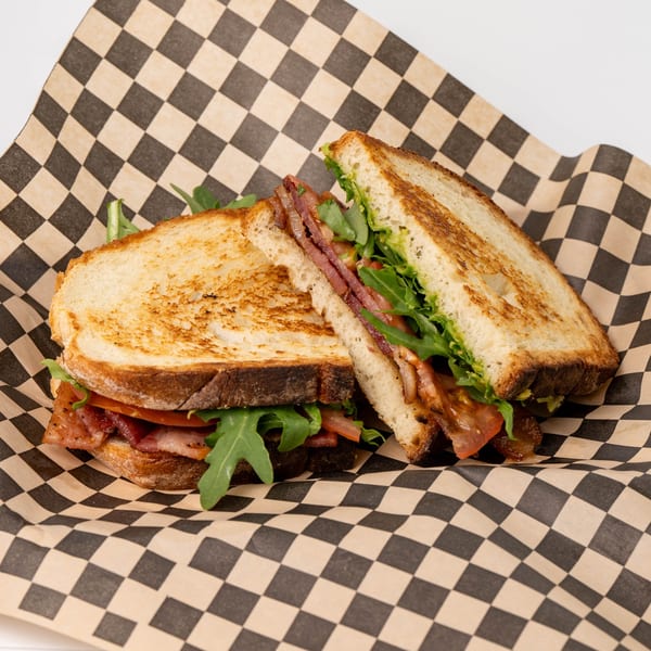 Toasted sandwich with bacon, arugula, and avocado on checkered paper from Jim & Patty's Coffee.