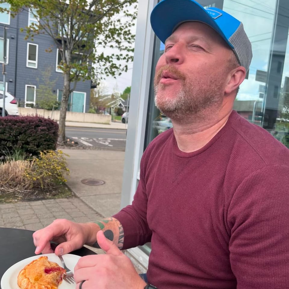Man in a blue hat and maroon shirt enjoying a pastry at an outdoor table.