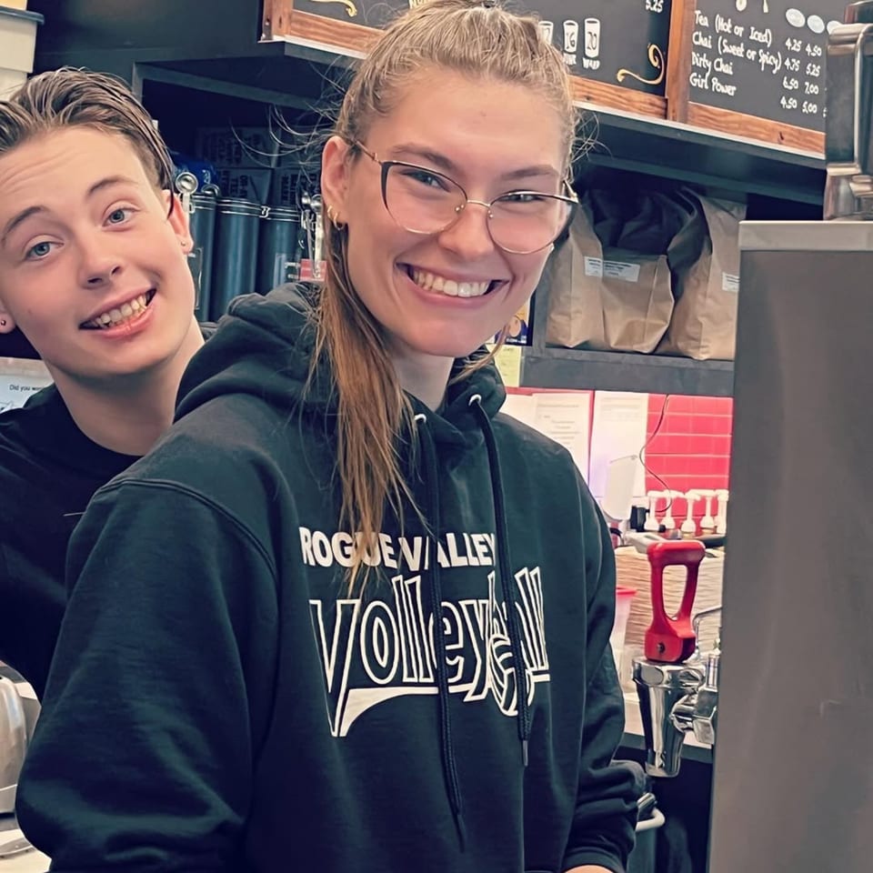 Two smiling baristas, one wearing glasses and a "Rogue Valley Volleyball" hoodie, behind the counter.