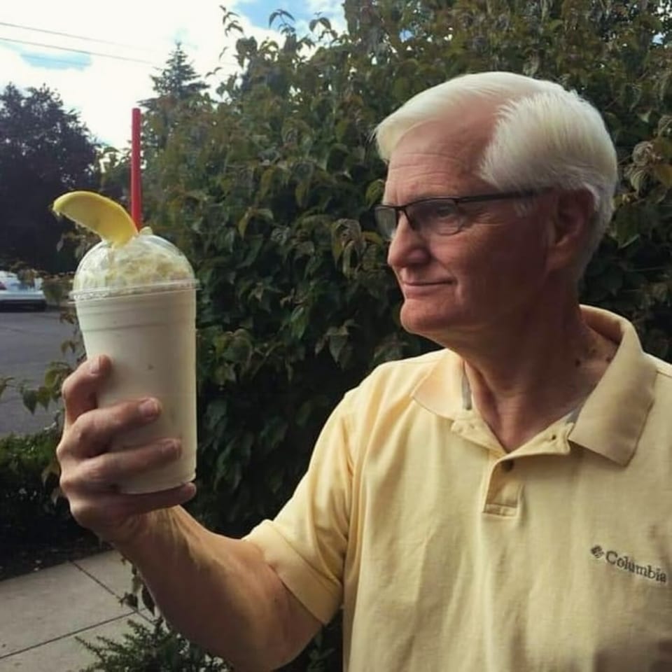 A man in a yellow polo shirt holds up a Jim & Patty's Coffee banana smoothie outdoors.