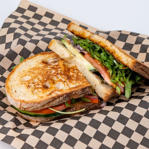 Grilled sandwich with arugula, tomatoes, cucumbers, and onions on checkered paper.